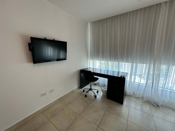 A modern office space with a flat-screen TV mounted on the wall, a black desk with a chair, and a large window with sheer curtains. - Estrella Federal I 2 Bedroom Apt in Costa del Este (Panama City)
