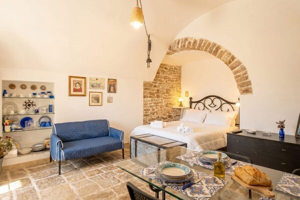 A cozy room with a bed, a blue sofa, a glass table with plates, and a stone wall. - Corte Fiorita – Intimate Suite in the historic center (Bitonto)
