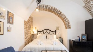 A bedroom with a stone arch, a bed with white linens, a nightstand, a dresser, and a blue sofa. - Corte Fiorita – Intimate Suite in the historic center (Bitonto)
