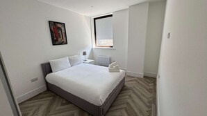 A modern hotel room with a large bed, a bedside table, a lamp, and a window with blinds. - Modern 2-Bedroom Apartment  in the Heart of Shoreditch (London)