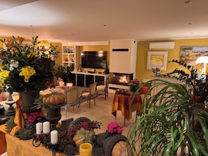 A cozy living room with a fireplace, a television, and various decorative plants and pumpkins.