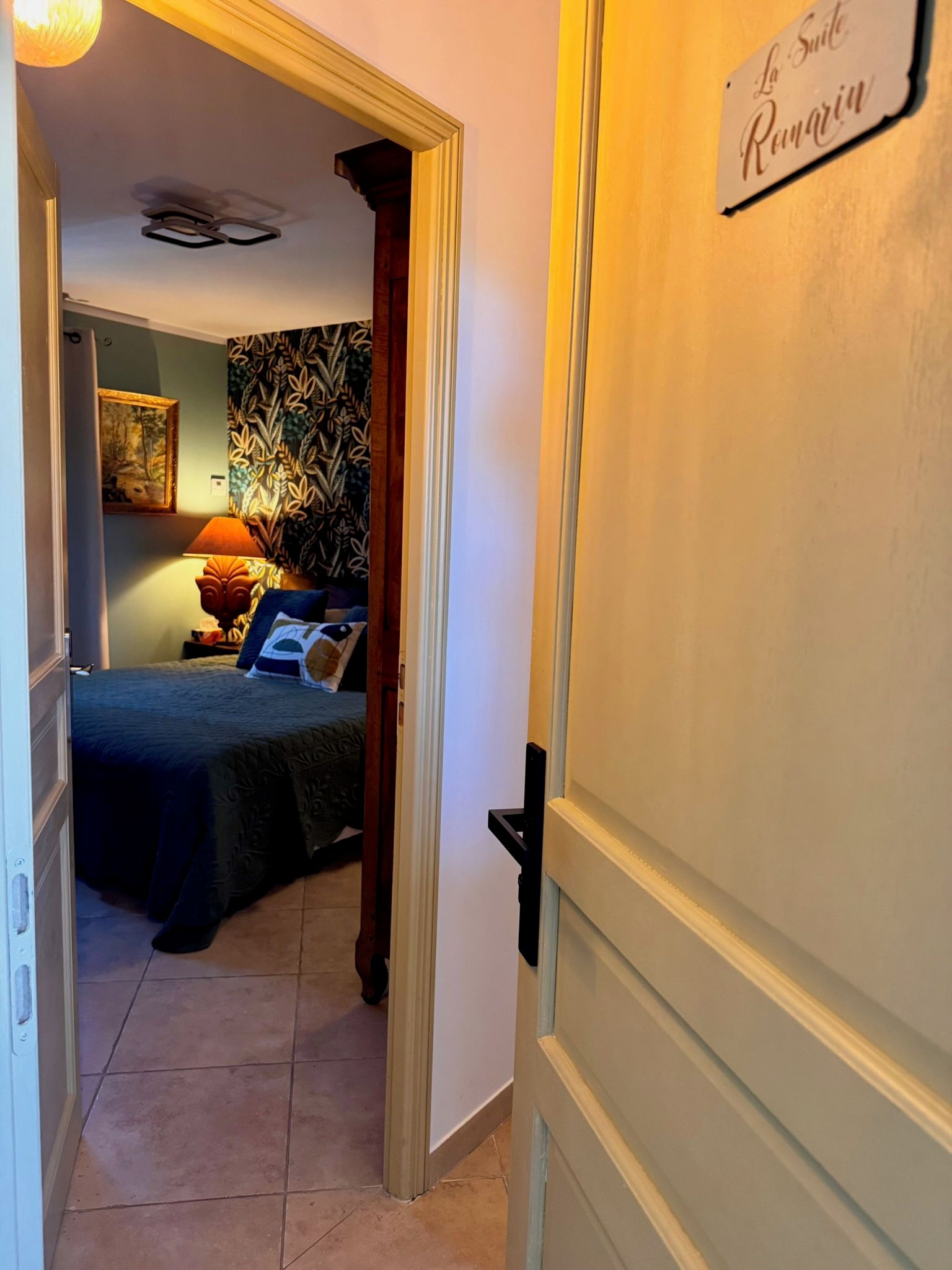 Een hotelkamer met een bed, nachtkastje, lamp en een muur met een bladmotief.
