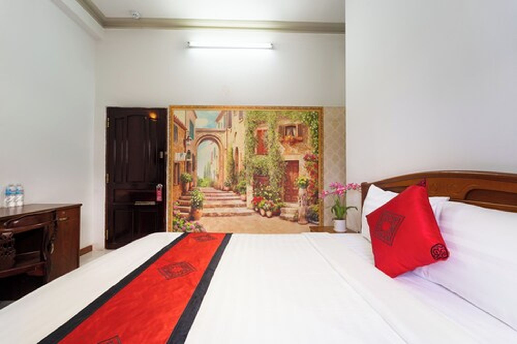 A hotel room with a bed, a wooden headboard, a red pillow, a white bedspread, and a painting of a street scene on the wall.