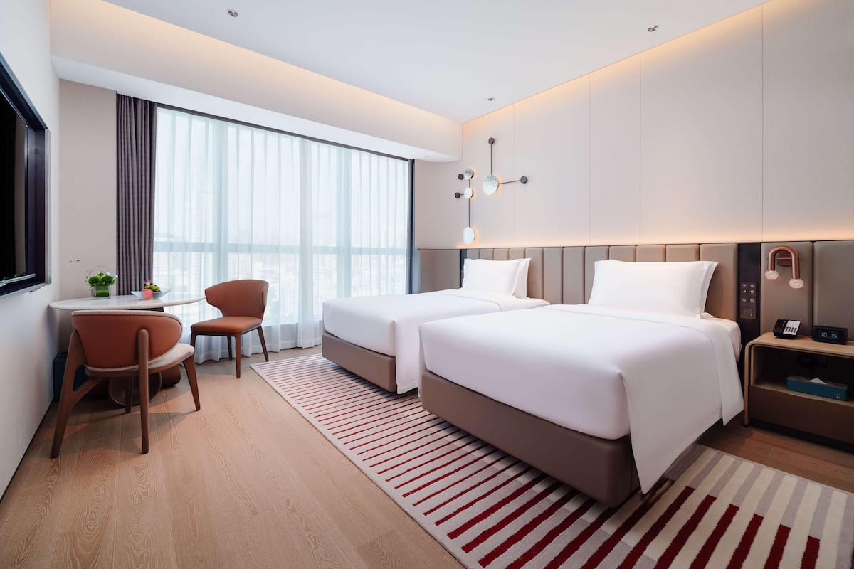 Una habitación de hotel moderna con dos camas, un escritorio, una silla y un amplio ventanal con cortinas.