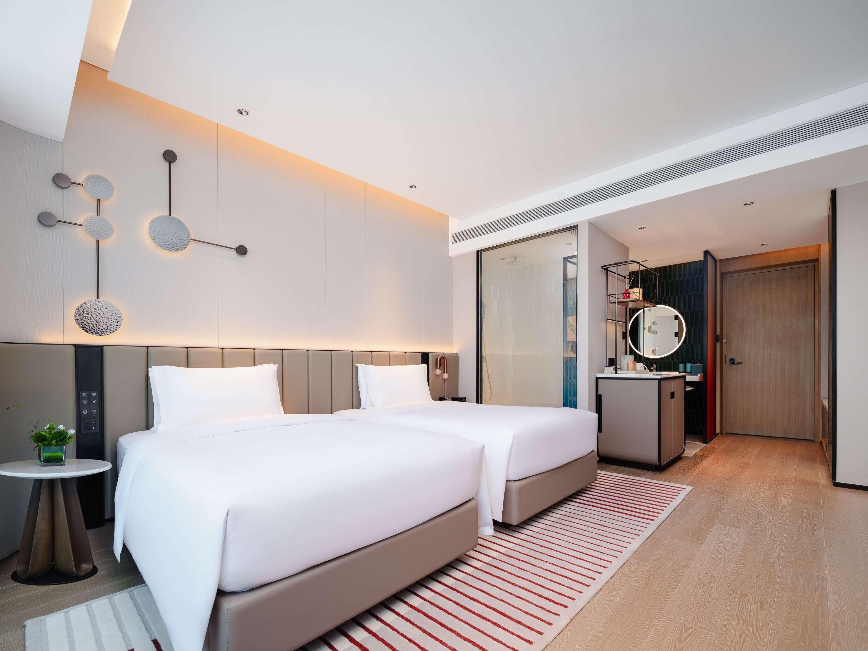 A modern hotel room with two beds, a small table with a plant, a mirror, and a bathroom with a shower.