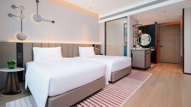 A modern hotel room with two beds, a small table with a plant, a mirror, and a bathroom with a shower. - Novotel Xiamen Downtown (Xiamen)