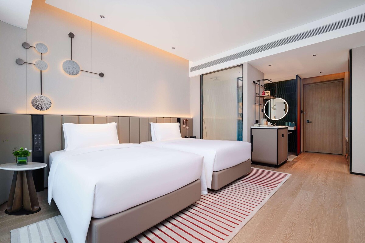 A modern hotel room with two beds, a small table with a plant, a mirror, and a bathroom with a shower.