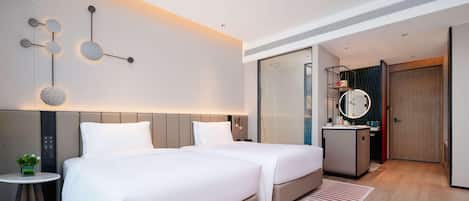 Quarto de hotel moderno com duas camas, uma mesinha com uma planta, um espelho e um banheiro com chuveiro.