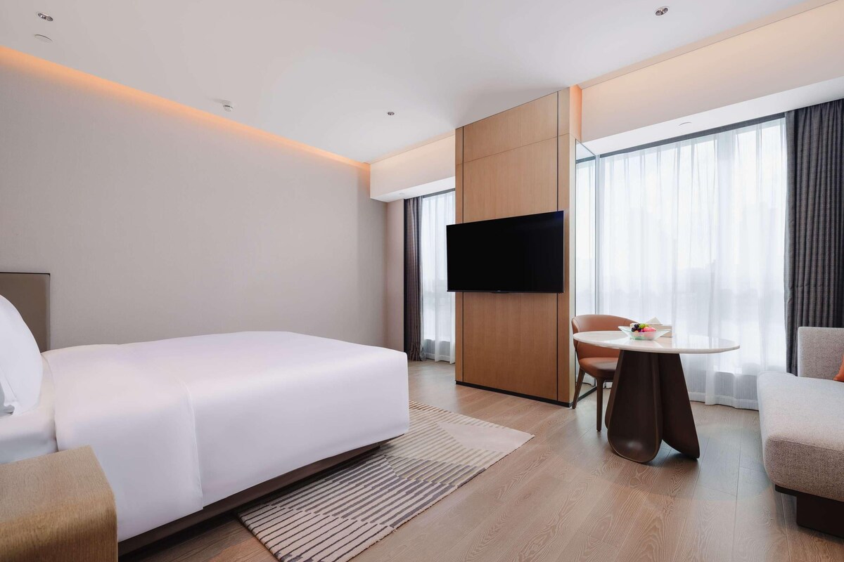 Una habitación de hotel moderna con una cama grande, un televisor de pantalla plana montado en un panel de madera, una pequeña mesa redonda con una silla y ventanas amplias con cortinas translúcidas.