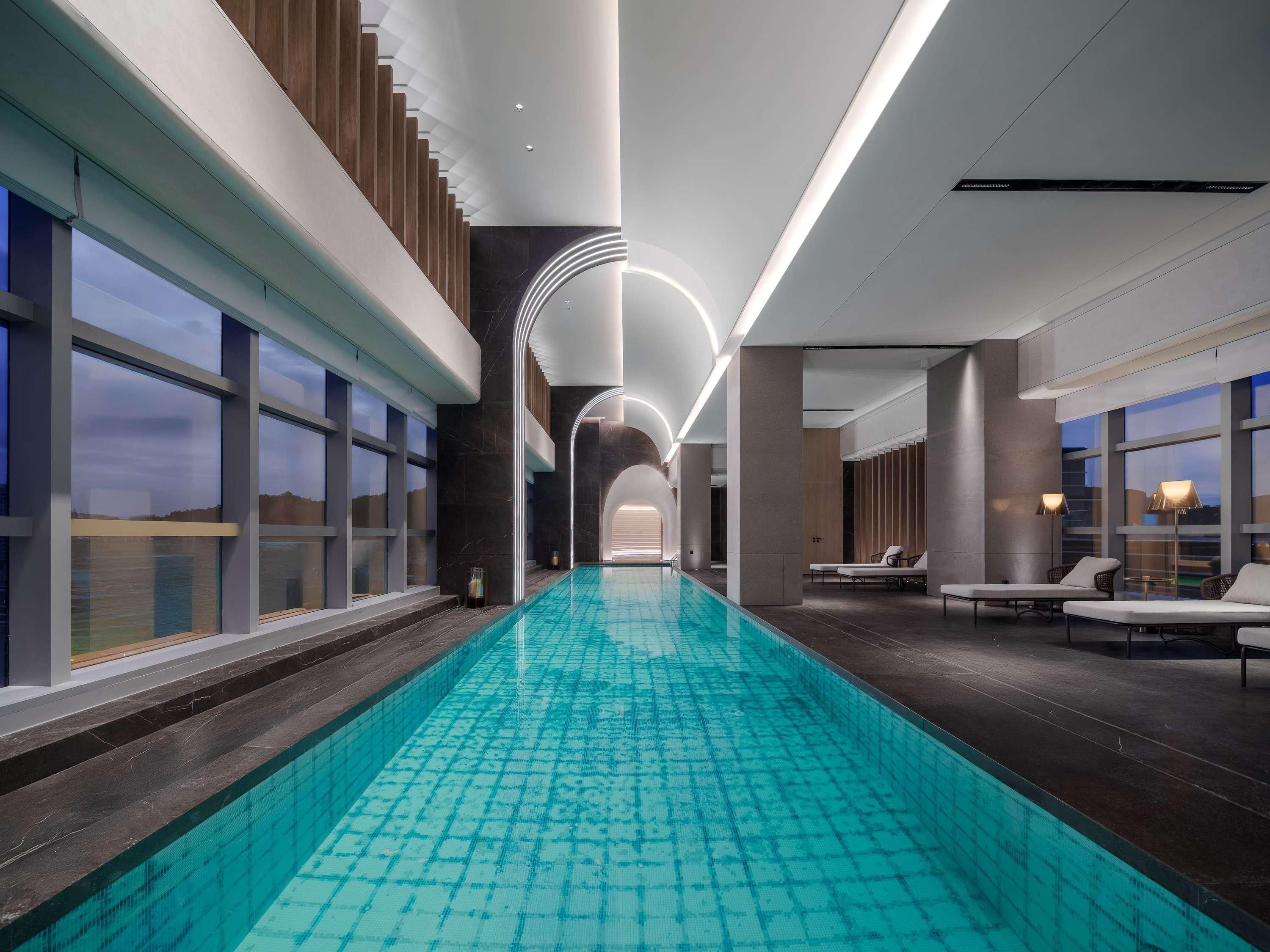 A modern indoor swimming pool with a clear view of the sky through large windows, a long poolside area with lounge chairs, and a contemporary design with clean lines and minimalistic decor.