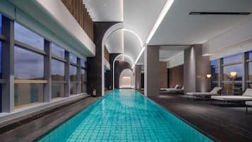 A modern indoor swimming pool with a clear view of the sky through large windows, a long poolside area with lounge chairs, and a contemporary design with clean lines and minimalistic decor.