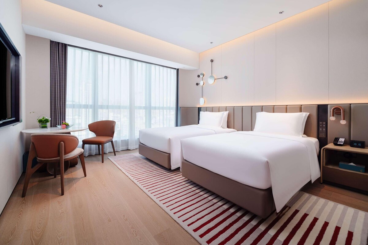 A modern hotel room with two beds, a desk, a chair, and a large window with curtains.