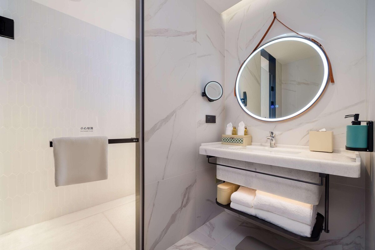 A modern bathroom with a glass shower door, a marble wall, a round mirror with a light, a sink with a towel rack, and a soap dispenser.