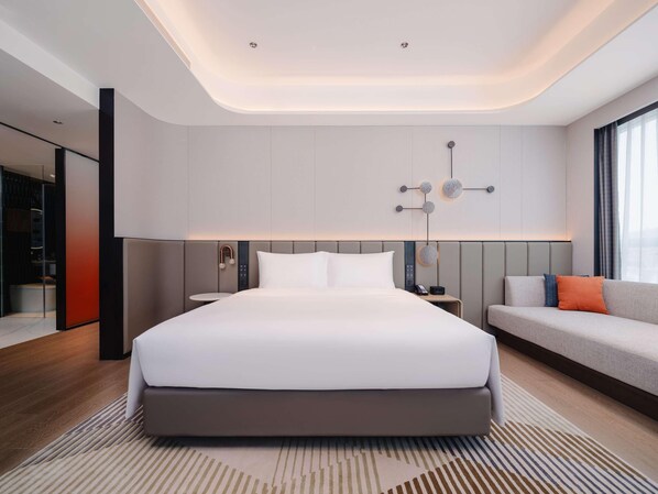 A modern hotel room with a large bed, a sofa, a bedside table, and a wall-mounted light fixture.