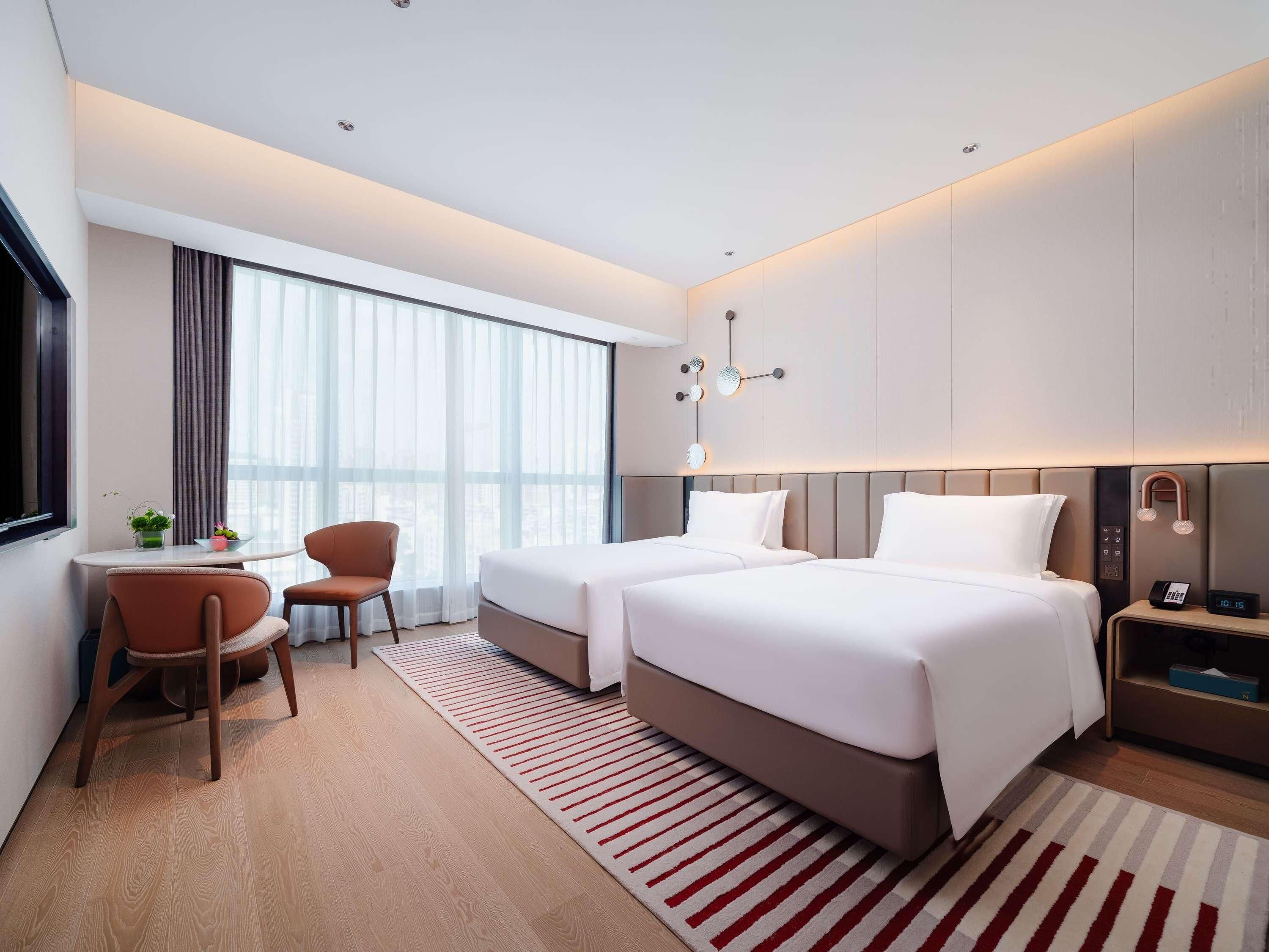A modern hotel room with two beds, a desk, a chair, and a large window with curtains.