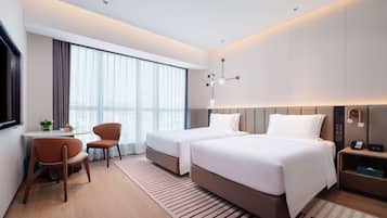 A modern hotel room with two beds, a desk, a chair, and a large window with curtains.