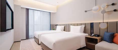 A modern hotel room with two beds, a large window, a flat-screen TV, and a bedside table with a phone and alarm clock.