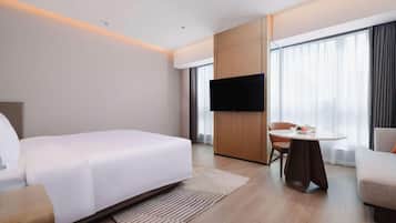 A modern hotel room with a large bed, a flat-screen TV mounted on a wooden panel, a small round table with a chair, and large windows with sheer curtains.