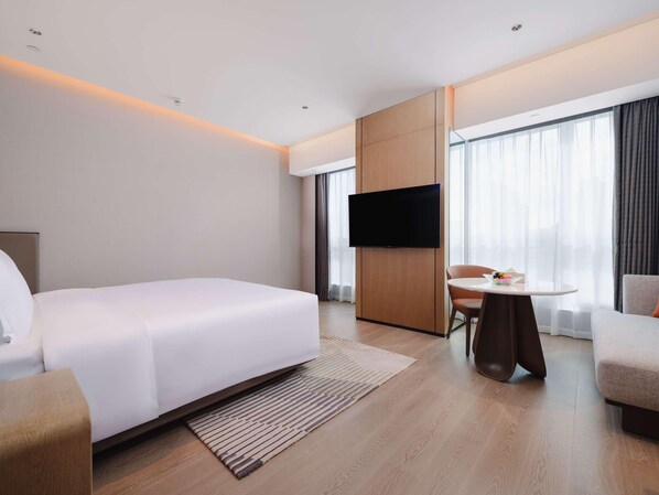 A modern hotel room with a large bed, a flat-screen TV mounted on a wooden panel, a small round table with a chair, and large windows with sheer curtains.