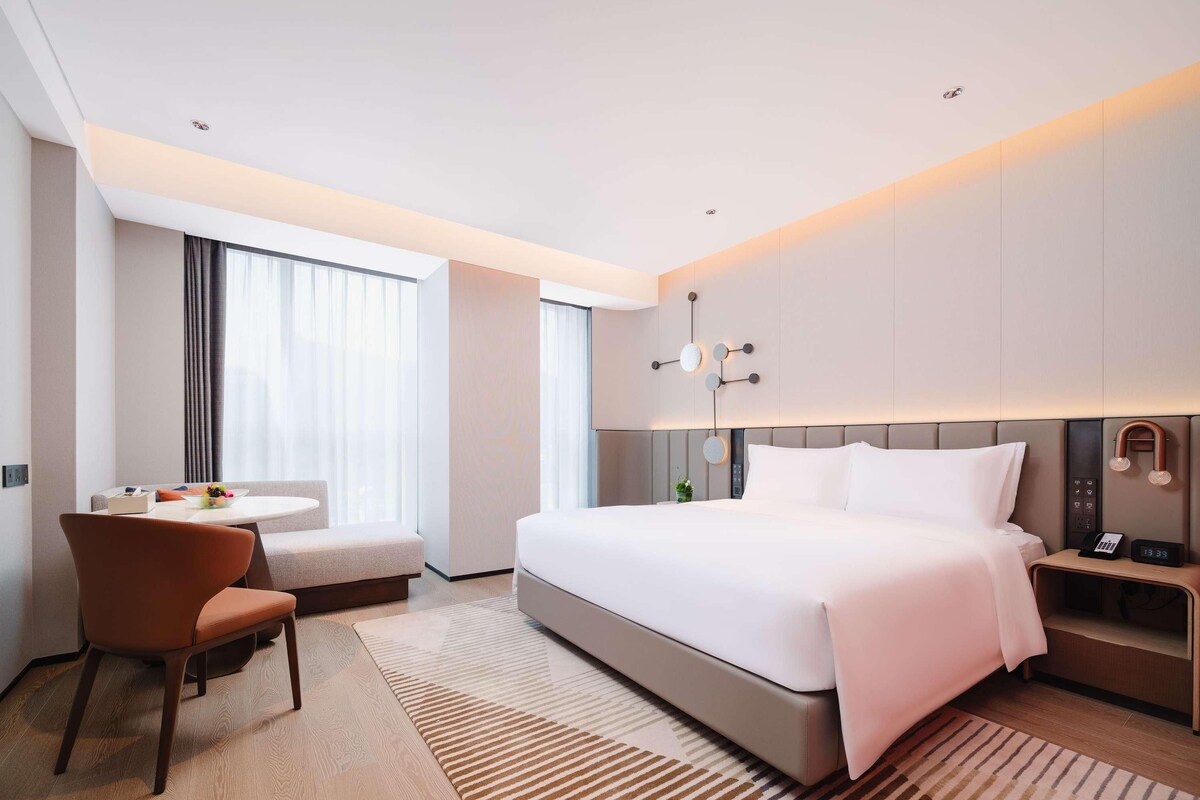Une chambre d’hôtel moderne avec un grand lit, un bureau avec une chaise, une petite table avec un saladier de fruits et une table de chevet avec une horloge.