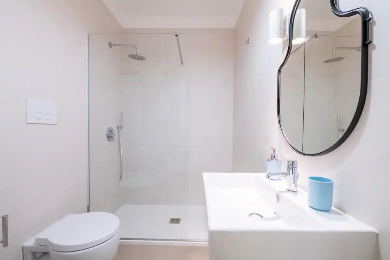 Ein modernes Badezimmer mit Duschkabine aus Glas, einer weißen Toilette, einem Waschbecken mit einer blauen Tasse und einem großen Spiegel.