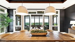 A dining area with a large wooden table, two pendant lights, and a view through large windows. - LUXURY HILLTOP ESTATE | 15 BEDS | POOL, SPA, SAUNA, FIRE PIT, POOL TABLE | 15 Mins to DT Austin (Austin)