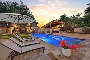 Uma piscina com espreguiçadeiras e guarda-sóis, rodeada por azulejos de pedra e vegetação.