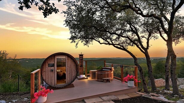 A wooden cabin with a large barrel-shaped door, a hot tub, and potted flowers on a deck overlooking a landscape. - LUXURY HILLTOP ESTATE | 15 BEDS | POOL, SPA, SAUNA, FIRE PIT, POOL TABLE | 15 Mins to DT Austin (Austin)