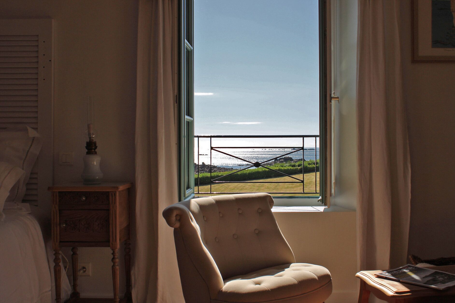Ein Zimmer mit großem Fenster und Blick auf eine Wiese und Wasser. Ein Sessel und ein kleiner Tisch mit Lampe stehen bereit.