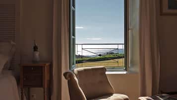 Ein Zimmer mit großem Fenster und Blick auf eine Wiese und Wasser. Ein Sessel und ein kleiner Tisch mit Lampe stehen bereit.