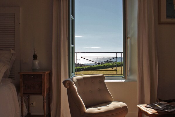 A room with a large window overlooking a grassy area and water. There is a chair and a small table with a lamp. - Chambres d'hôtes face à Bréhat (Ploubazlanec)