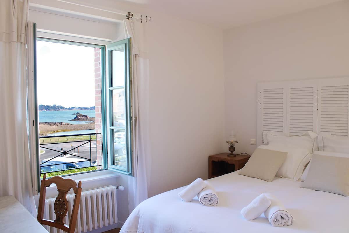Ein Schlafzimmer mit Bett, Nachttisch, Sessel, Fenster mit Meerblick und Heizkörper.