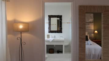 Ein Badezimmer im Hotel mit einem weißen Waschbecken, einem Spiegel und einer Toilette. Ein Stehlampen und ein Bett mit weißer Decke sind ebenfalls vorhanden.