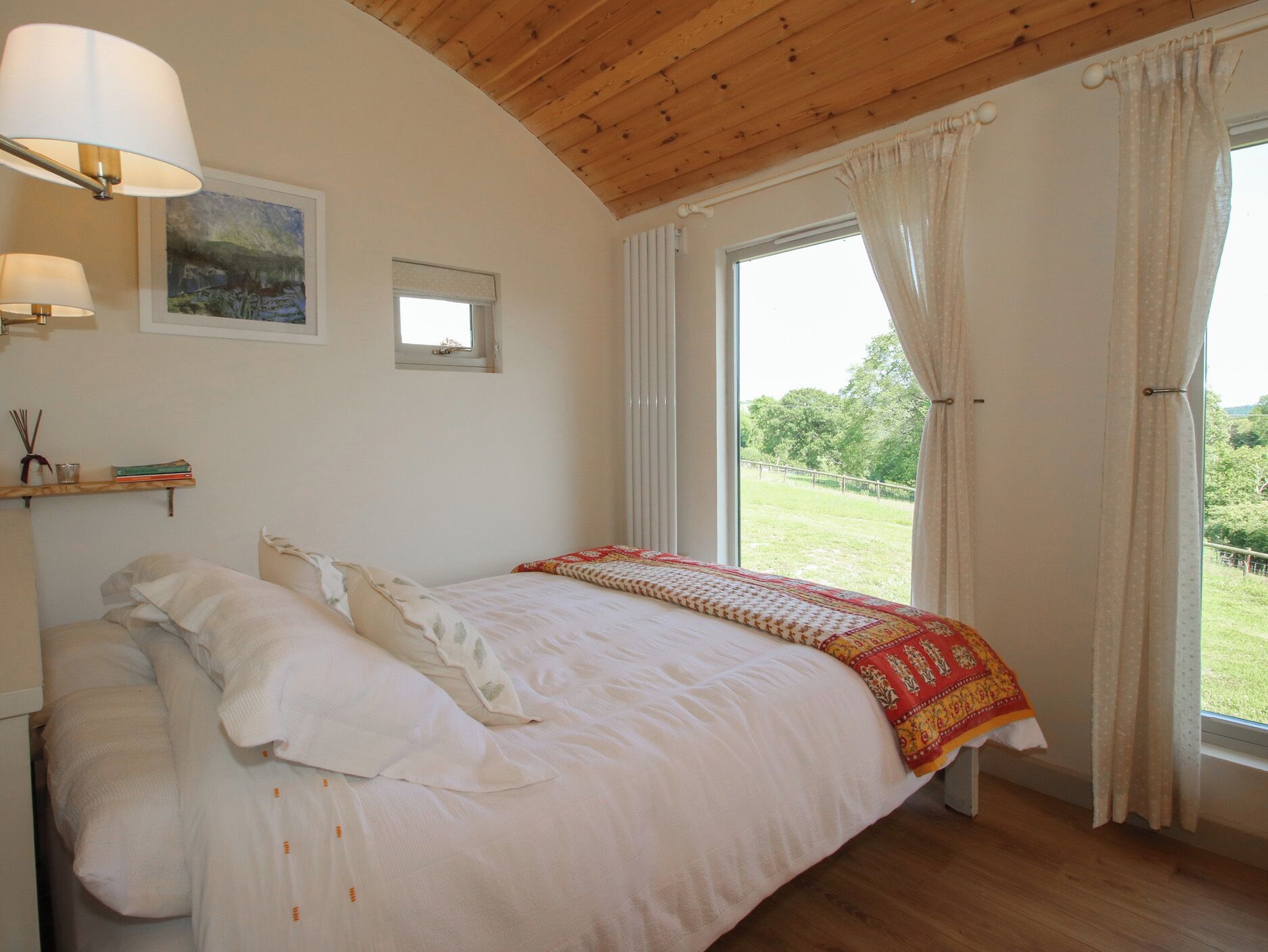 A bedroom with a wooden ceiling, a bed with white bedding, a window with curtains, and a painting on the wall.