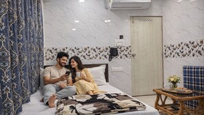 A bedroom with a bed, a bedside table with a phone, a door, and a small table with a bowl and flowers. - Hotel Relax Kurla  (Mumbai)