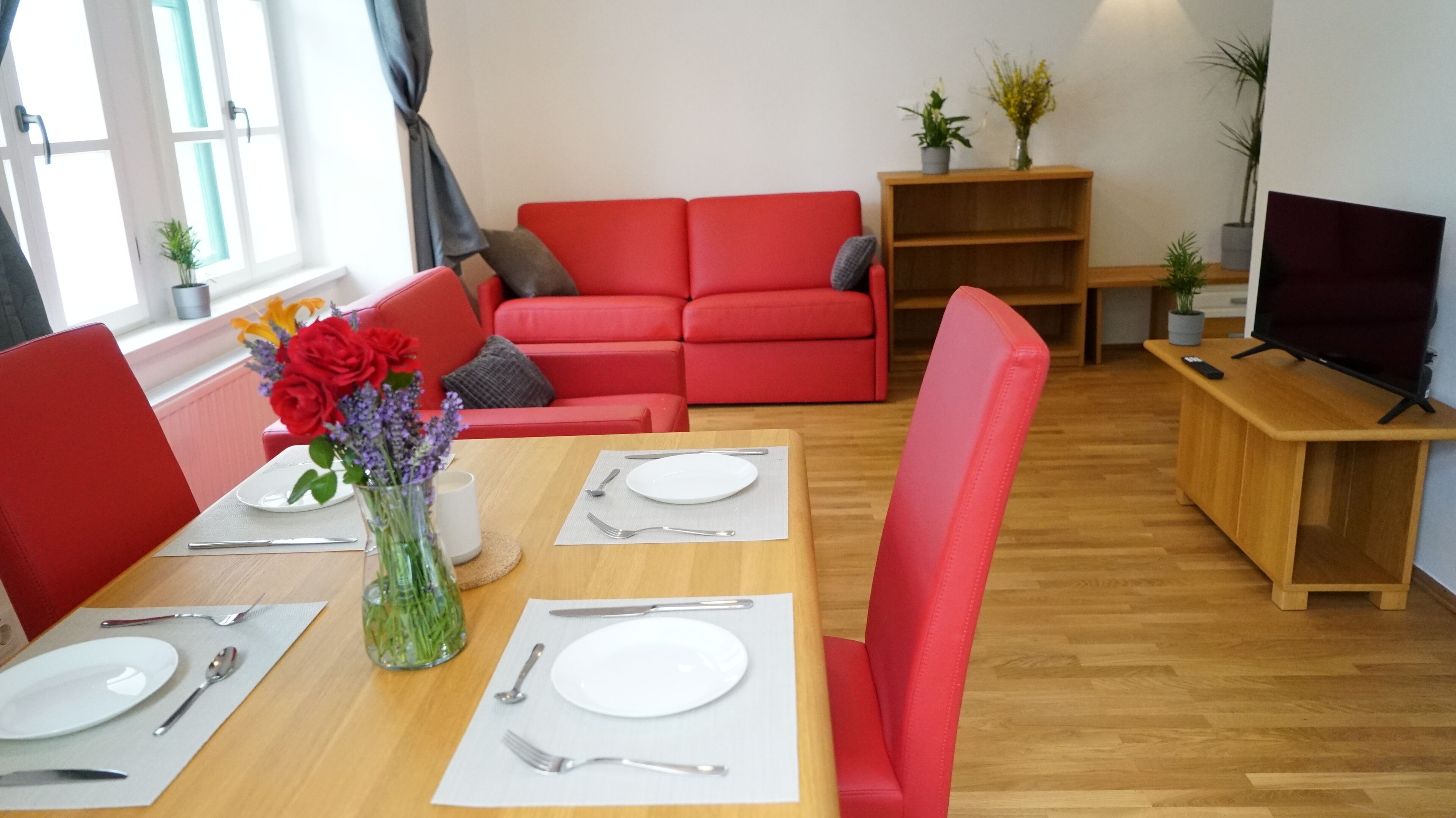 A dining area with a wooden table set for two, red chairs, and a vase with flowers.