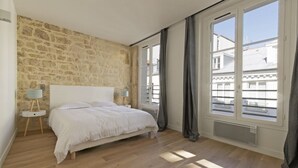 Una camera da letto con parete in pietra, un letto, un comodino con lampada e ampie finestre con tende.
