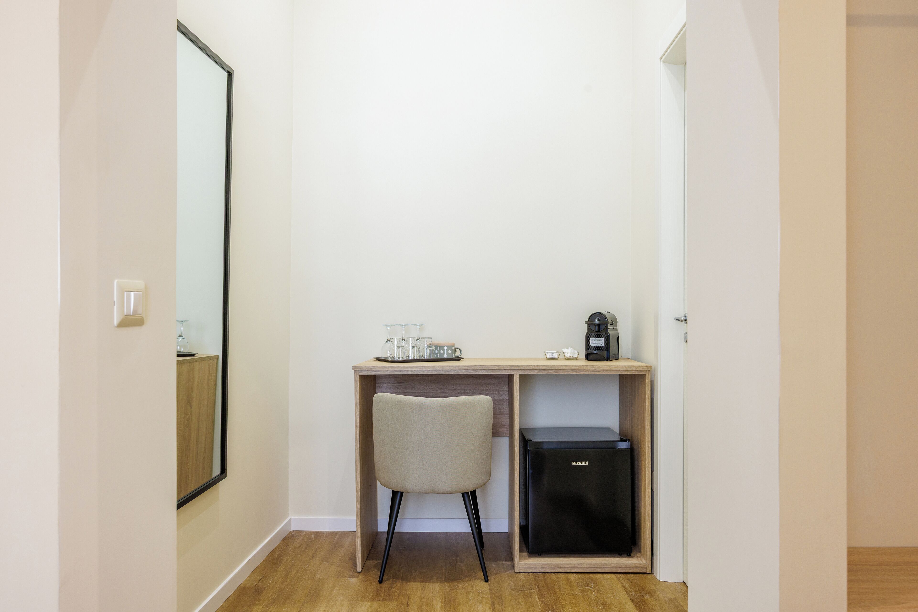 A small, well-lit room with a wooden desk, a chair, a coffee maker, and a mini-fridge.