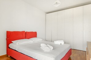 A bed with white bedding and red headboard, a white wall, and a wooden floor.