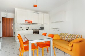 A modern kitchen with white cabinets, a dining table with orange chairs, and a yellow sofa with striped pillows.