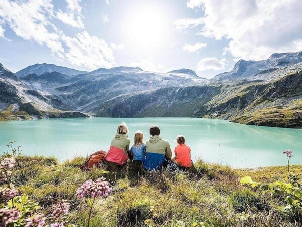 Eine Gruppe von Menschen sitzt auf einer Wiese mit Blick auf einen See, im Hintergrund Berge.