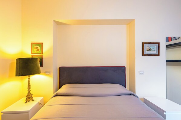 Een net opgemaakt bed met een grijs hoofdbord, een bedlampje en twee ingelijste foto's aan de muur.