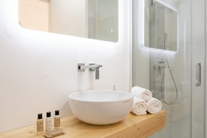 A modern bathroom with a white vessel sink, a wooden vanity, a glass shower enclosure, and neatly rolled towels.