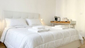A neatly made bed with white bedding and towels, a bedside table with a lamp, and a small wooden nightstand with a cup and a box.