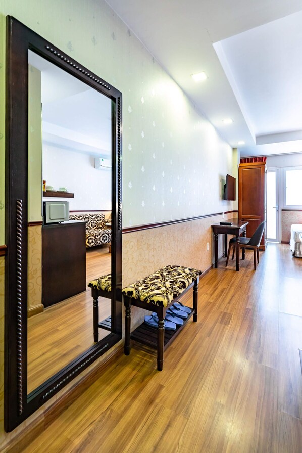 A hotel room with a wooden floor, a large mirror, a bench, a small table with a chair, and a television on a stand. - Dancentre Luxury Apartment Near SECC (Ho Chi Minh City)
