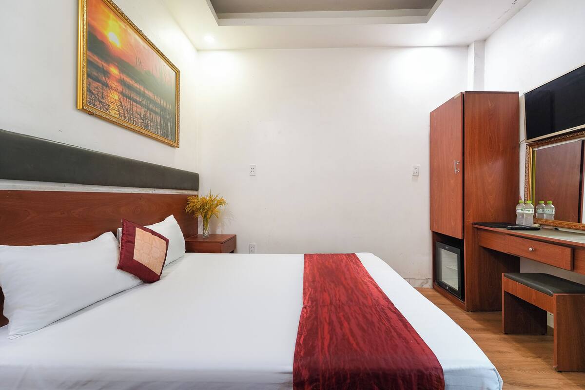 A neatly made bed with white and red bedding, a wooden headboard, a painting of a sunset, a small table with a vase of flowers, and a wooden wardrobe with a mirror.