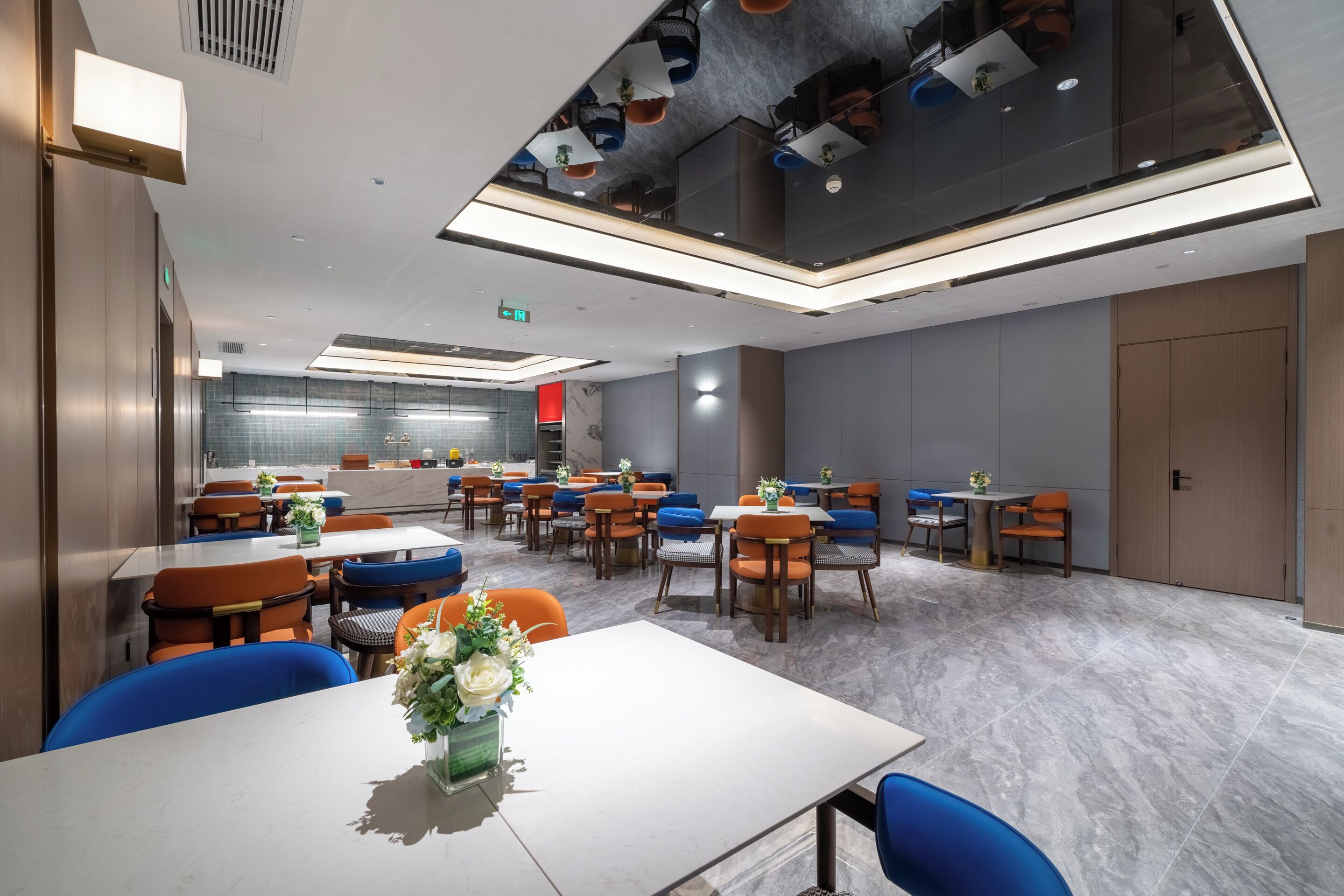Un espace de restauration moderne, doté de plusieurs tables et chaises, avec une cuisine ouverte visible au fond et un plafond pourvu d’un éclairage encastré.