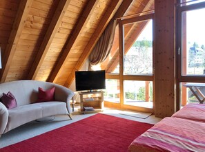 A room with a wooden ceiling, a sofa, a TV on a stand, a bed, and large windows.