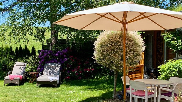 A garden with a patio area, including a table and chairs, a large umbrella, and various plants and flowers.