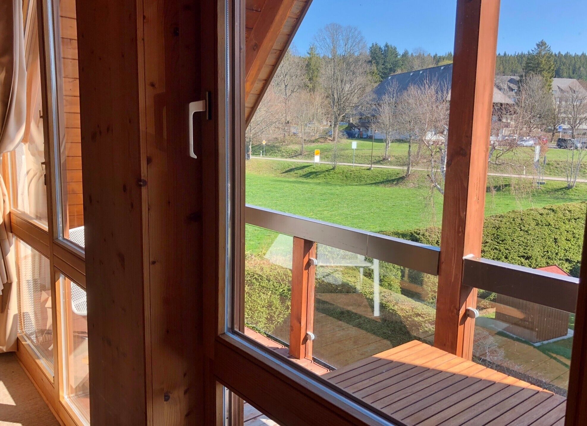 Ein raumhohes Holzfenster mit Blick auf eine grüne Landschaft, eine Holzterrasse und einen klaren Himmel.